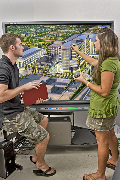 Woman stands to the right, touches giant screen with writing tool while male sits to the left holding a book and watching.