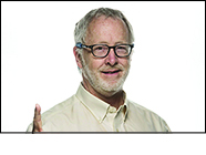 Bearded man in glasses, smiles and poses with index finger extended out to represent the number one, wears pale yellow shirt in front of a white background.