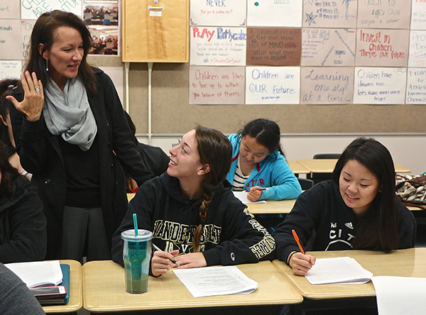 Three young ladies sit at desks, writing in packets wearing black hoodies, while another more mature woman, also wearing black, stands over them, talking.