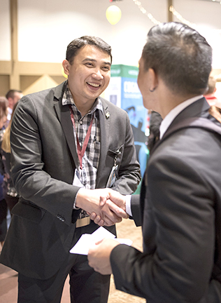 Young man, dressed in a gray suit smiles while he shakes hands with another young man whose back is to the camera on the right side of the image.