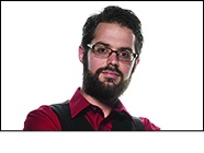 Bearded man, wearing rectilinear framed glasses, dressed in a red shirt and black vest poses in front of a white background.