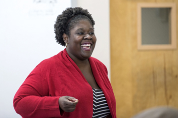 A woman, wearing a bright red sweater over a black and white striped garment, smiles warmly while holding something small in her right hand; her arm folded in front of her body.