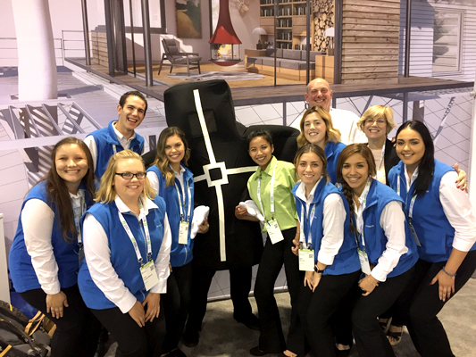 Eight individuals wearing blue vests and lanyards pose in front of a curtain emblazoned with an artist depiction of both the outside and inside of a room space.