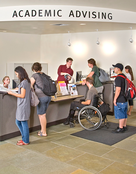 Students gather at Academic Advising counter. Two folks get help from desk attendants. One stands at the check-in computer while two others wait in line.