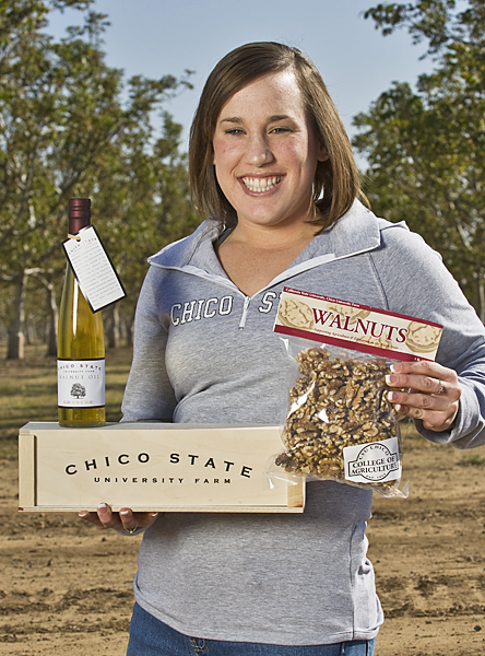 Brunette woman wearing long-sleeved gray Chico State sports shirt smiles, center frame, holding a bag of walnuts in her left hand and balancing a wine bottle on top of a custom wine box displaying "Chico State University Farm."