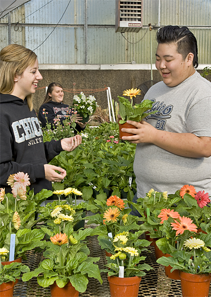 Two students smile and gaze upon a flower inside of a nursery. Woman holds flowers in background.