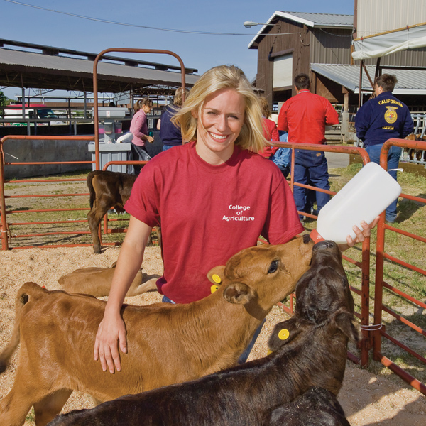 Smiling woman in red shirt holds milk bottle in left hand feeding calves; right hand rests on calf.