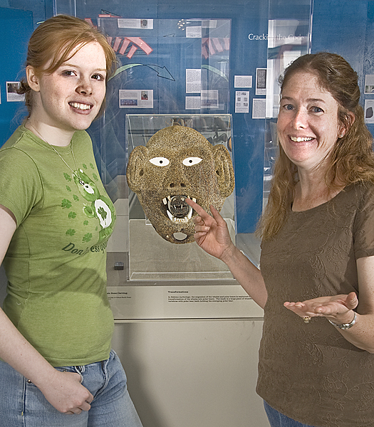 Two women stand in front of a mask that is part of a display.