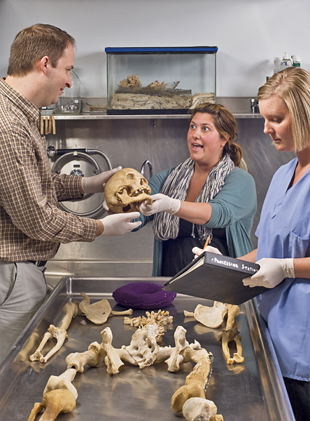 Woman (center) hands human skull to man while another woman holds and writes on a binder labeled "Processing Statistics."