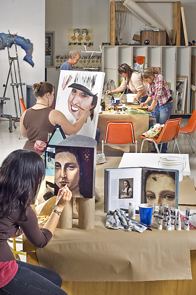 Art studio, two women on left paint portraits, one sits, three people stand, paint at table in back.