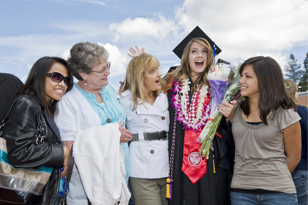 Five women stand interlocking arms behind them. The fourth woman wears graduation cap and gown, adorned with Hawaiian leis and holding a couple of flower bouquets. She smiles a big open-mouthed smile. The woman to her left shares the same smile wearing a double-breasted white jacket. Two women, one older, one younger, stand smiling and looking at the graduate, while a dark-haired woman stands to the grad's right.