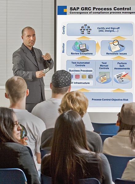 Man in grey suit holds remote and talks in front of presentation of "SAP GRC Process Control." The man gestures with his hands towards a group of people sitting in rows. In the back row, foreground of the photo, a dark-haired woman turns her head smiling in the direction of the woman sitting to her right. 