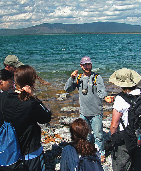 Along the shore of some body of water, a man holds a specimen of the natural inhabitants of the local ecosystem, and talks to a group gathered to listen.