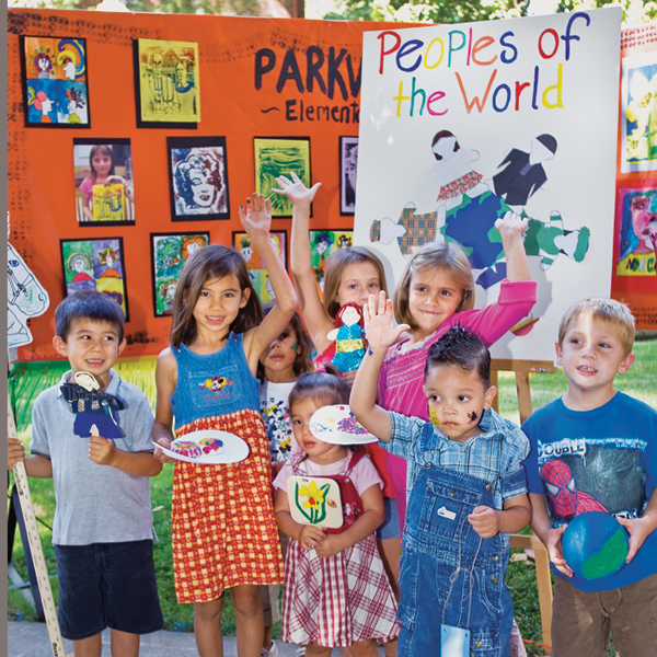 Smiling kids stand in front of artistic display, raising hands and holding crafts.