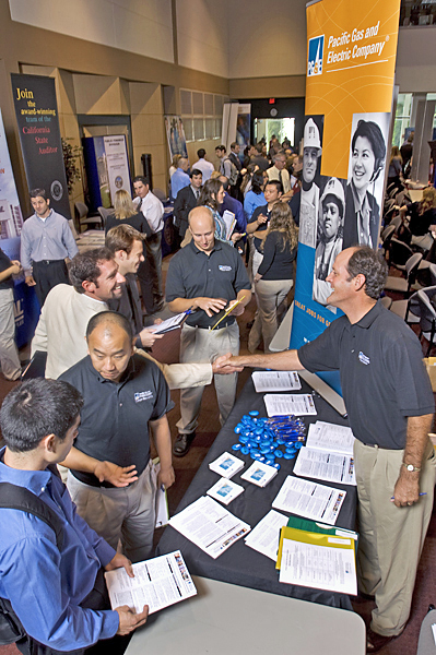 People gather at a business fair. In the foreground a student wearing blue shirt and backpack holding a sheet of paper speaking to a PG&E rep. In the middle-ground, an orange PG&E banner hangs above a different company rep shaking hands with another man. In the background, students, company reps, and employees mingle at a recruiting fair.
