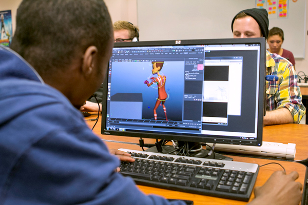 An over-the-shoulder shot, that peeks over the right shoulder of a man wearing a blue hooded sweatshirt as he sits in front of a computer.  On the monitor is an abstract animated human-like male.  Directional arrows and rings encircle the animated figure as the man at the computer maneuvers the mouse with his right hand.