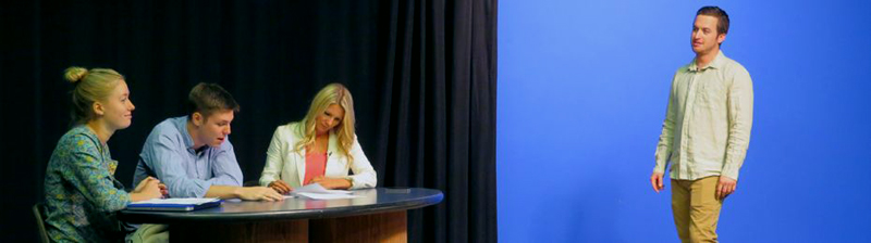 Photo is visually split.  The left side features three people sitting at an oval-shaped table against a black curtain.  A woman in green with her hair in a bun facing to the right.  A man in a blue shirt sits to the right of her speaking while looking down.  A woman in a pink shirt with white blazer sits at the end of the table holding papers. The right side features a man wearing a long-sleeved button down shirt and light brown pants standing in front of a blue screen.