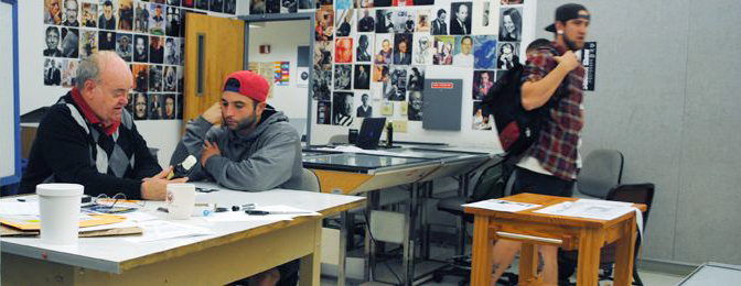 A man in a fetching argyle sweater sits at a table to the left of the image having a discussion with a man who wears his ball cap backwards.  Another man is blurred as he moves from right to left through the frame on the right .  The image is from the inside of a classroom facing out towards the door.  Portraits adorn the wall space on either side of the open door.