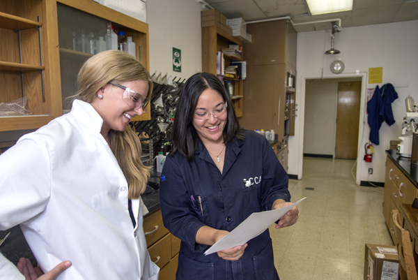 Two female students stand in a lab.  One student wears a white lab coat (left), while the other wears a navy blue lab coat.  Both wear safety glasses and are looking at a couple of papers that the student in navy is holding.