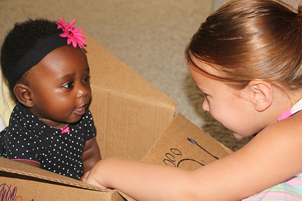 A baby sits in a box, fixated on another young girl smiling at her from outside the box.
