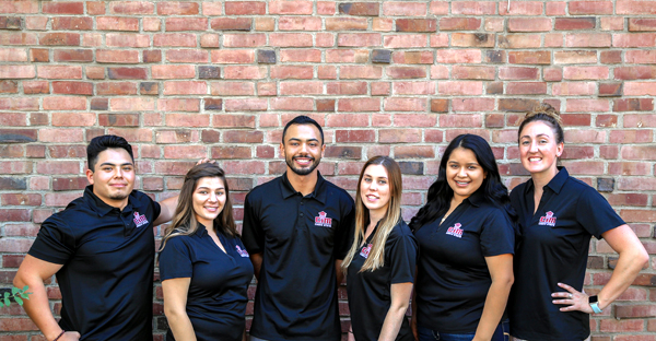 Six men and women, all wearing black nylon, short-sleeve, polo shirts with CIM printed in red letters with a white outline, smile in front of a brick wall on the Chico State campus.