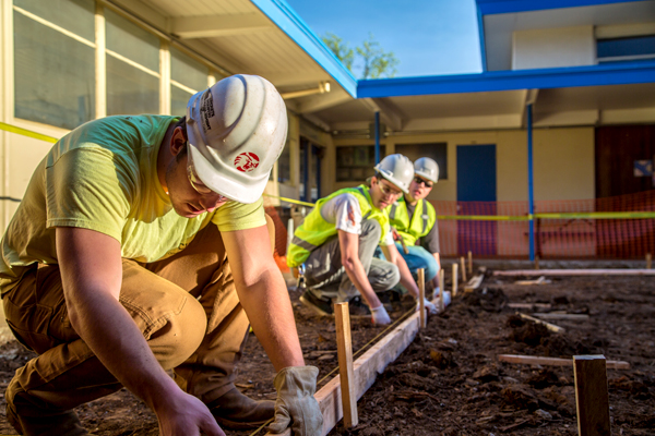 Three Chico State wearing white hardhats and bright, neon yellow vests, hold a wooden board in place in preparation of concrete pouring in the atrium at a local elementary school.