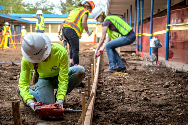 A woman with a hard hat, in a long-sleeved neon yellow shirt uses an electric screwdriver/drill at an angle parallel to the ground.  In the background, several CIM students in hard hats and vests work to prepare for concrete pouring.