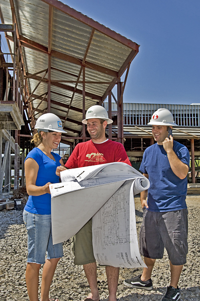 Three people wearing hardhats go over blueprints near construction zone. 