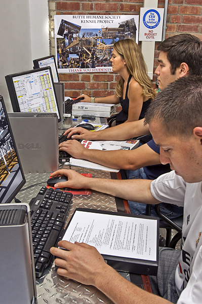 Three students sit at computers next to a poster thanking students for their work on the Butte Humane Society's Kennel Project.