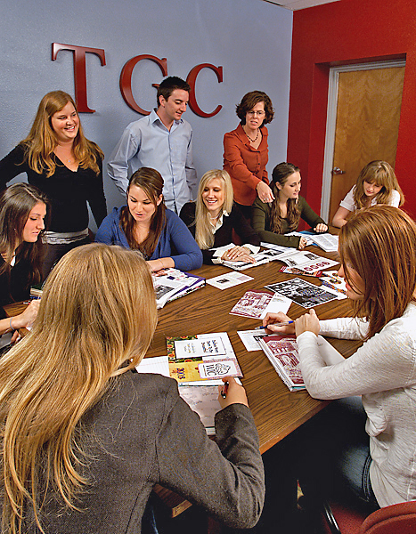 A group of women, and one man, convene around a large table reviewing and discussing brochures.