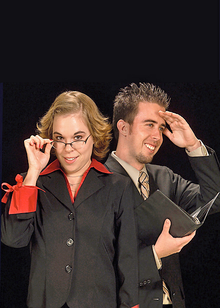 Man and woman pose against black background, dressed in business attire and smiling. Man salutes while woman looks over the top of her glasses.