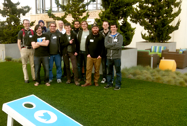 A group of men wearing name tags stands on lawn made of artificial turf.  Evergreen trees in planters behind the men disguise the building in the background. Off to the right of the image is an arrangement of patio furniture.  In the foreground is a cornhole board with the Twitter logo emblazoned on face of the board.