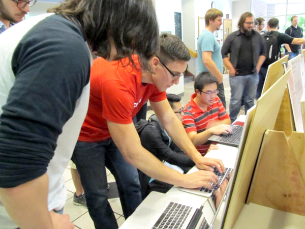 The photo is of a group of men working on a bank of laptop computers on a long table. The table is positioned from bottom center moving up and to the right out of frame.  The focus is on a pair of men in the foreground.  One man in a short sleeved red polo shirt leans down and works on one laptop, facing from left to right.  The other man, leans in over him, with his back to the viewer, long hair obscuring his face.
