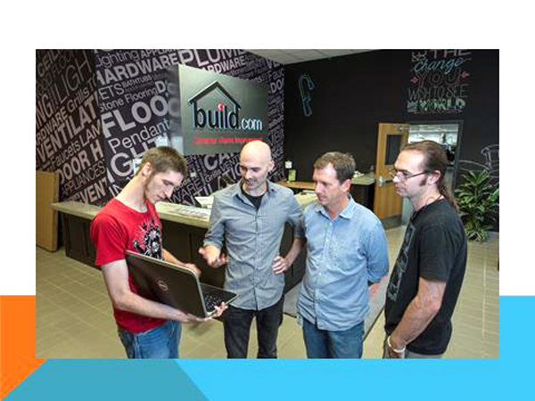 Four men stand in a line inside Build.com's lobby staring at a laptop computer.  One man in a red t-shirt, stands to the far left holding the laptop.  The man second from the left, gestures with his right palm up.  The other two men stand, one with arms behind his back and the other with arms at his side, watching intently.