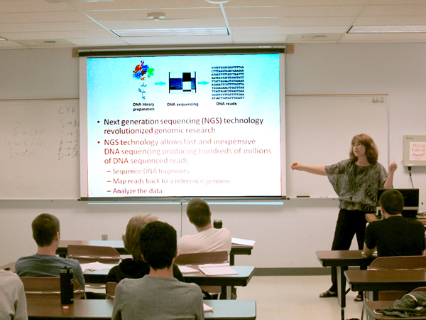 In a classroom setting, a woman stands at the far right, with her right arm extended out toward a screen facing the viewer.  Several students sit in desks with backs to the viewer.  On the screen is an information graphic discussing next generation sequencing technology.