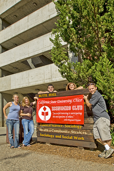 Students surround Butte Hall sign that is covered up by an Economics Club sign being held by two of the five people.