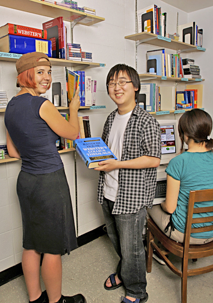 Woman pulls book off of shelf standing next to a man holding a dictionary while another woman sits at a computer behind him.