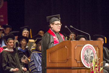 Graduate, dressed in black cap and gown, with shiny, bright red sash draping around shoulders, stands at a podium addressing an audience that is out of frame.  A group of other graduates and faculty sit in the background.