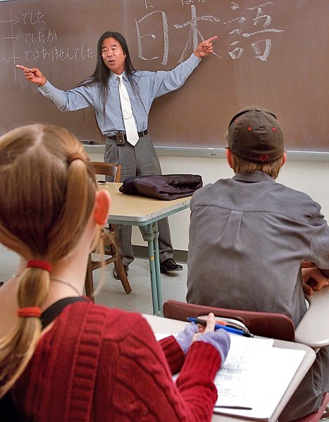 Man with really long hair stands in front of class with his arms spread and fingers pointing in opposite directions. There are Japanese glyphs on the chalk board behind him.