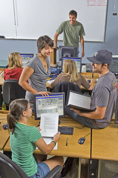 Three students talk with each other around a computer; one man holds binder, one lady touches monitor and one woman holds a packet of papers.