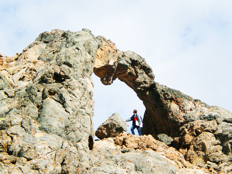 Man wearing a backpack stands under a naturally occurring rock arch.