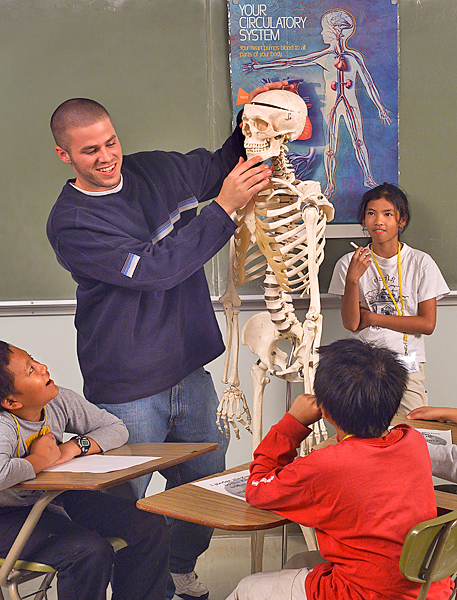 Adult man stands near two boys' desks holding a skeleton by the skull with a poster on the blackboard that reads "Your Circulatory System."