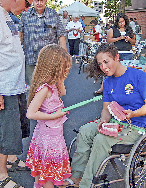 A little girl holds a large toothbrush ready to brush a large set of teeth that a woman in a wheel chair holds in her lap.