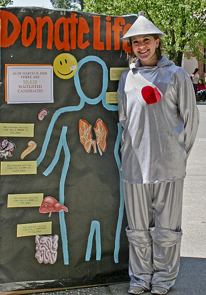 Woman dressed as the Tin Man with a heart tacked on stands smiling next to a poster with an outline of a human form showing organ cut-outs and the words "Donate Life" across top.