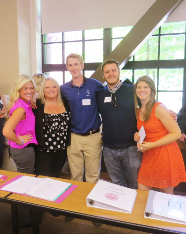 Five students stand arm-in-arm in a line in front big women and behind a long table with three binders on it.  From left to right: A blond woman wearing a bright pink vest and gray slacks embraces another blond woman wearing a black top with white polka dots. She hugs a man in a blue polo shirt with his arm around another man wearing a navy blue v-neck sweater and embracing a woman wearing a bright orange dress.