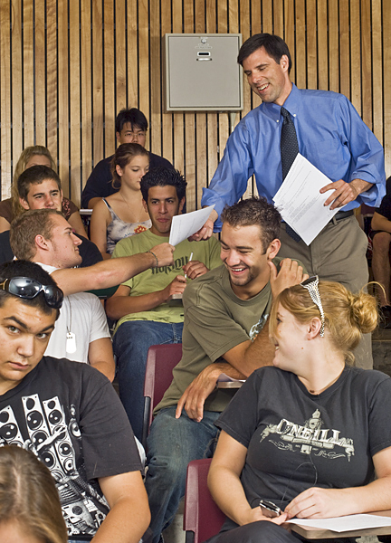 Professor stands in a stadium-seated classroom and smiles as he hands a student a packet of paper.