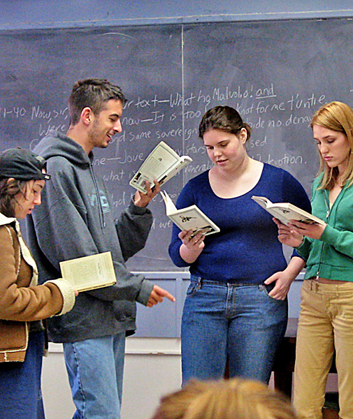 Four students, each holding a copy of the same book, stand together near a chalk board. Man points one finger while reading. The three other students follow along in their books, listening.