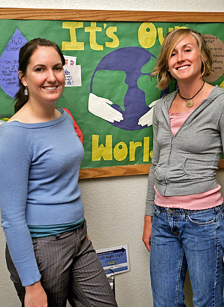 Two women smile standing in front of a display board with yellow words spelling "It's our world" juxtaposed against a green background with an abstract Earth being held by a pair of hands.