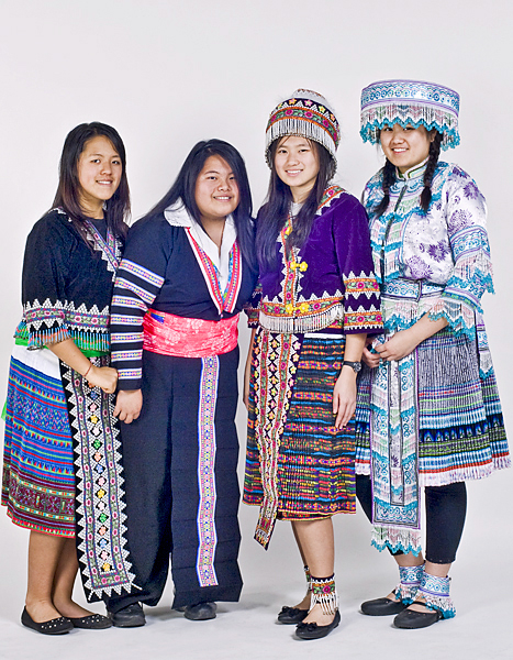 Four women, all wearing colorful traditional/cultural clothing, pose together and smile. Two of the women adorn fancifully decorated hats that match their outfits.