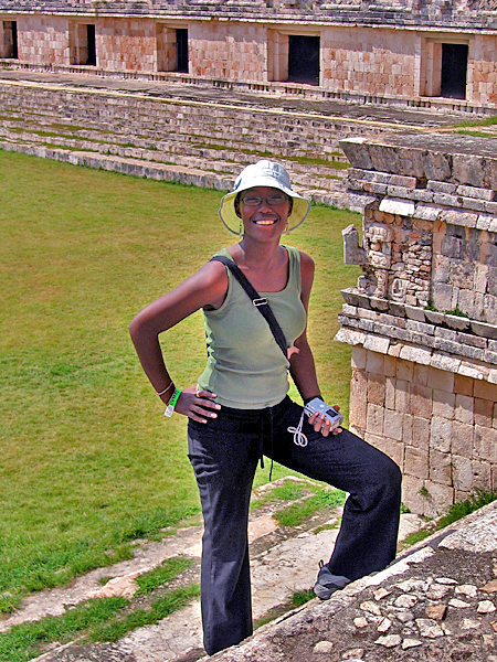A woman wearing a fishing hat and carrying a camera, amid ascension of the stairs of an ancient ruin, pauses to pose and smile for a picture.